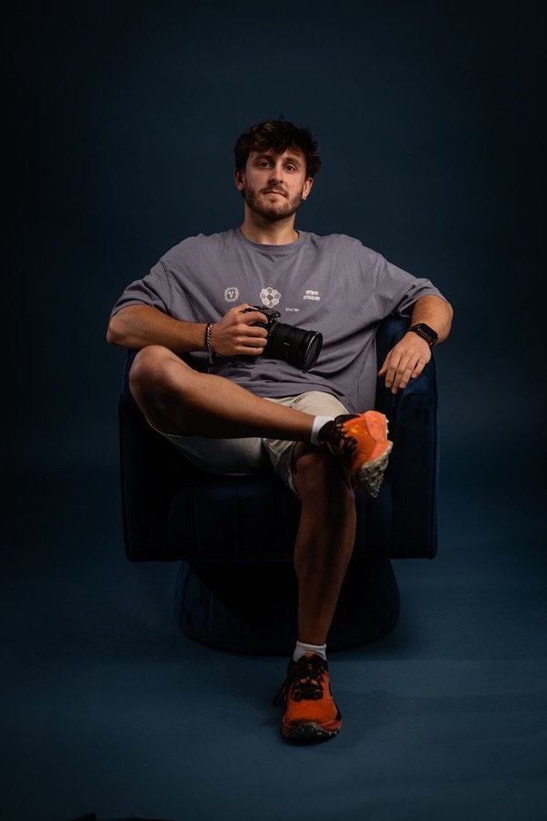 Retrato de Jan Castany sentado con una cámara en la mano sobre un fondo azul.
